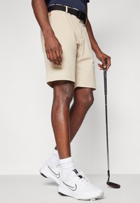 Bežiniai golfo šortai su tiesiu kirpimu, juodas diržas ir kišenės detalė. Dėvimi su baltais Nike golfo batais ir juodu golfu rankoje.
