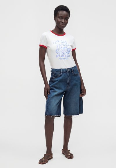 Γυναίκα που φοράει λευκό T-shirt με ρίγες, με γραφικό πίτσας και κείμενο, σε συνδυασμό με τζιν σορτς μέχρι το γόνατο και καφέ σανδάλια, στέκεται σε χαλαρή στάση.