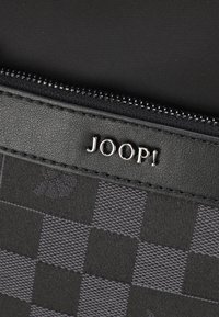Pochette nera testurizzata con chiusura lampo, caratterizzata da un motivo a scacchi e un logo metallico "JOOP!" su una superficie liscia.