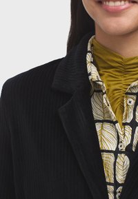 Veste en velours côtelé noir avec un col notched, superposée sur un turtleneck jaune froncé et une chemise à motif feuillage noir et blanc.