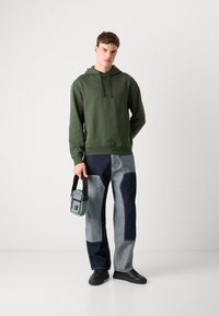 Felpa verde oliva con cappuccio e coulisse, abbinata a pantaloni di jeans a patchwork in tonalità chiare e scure. Tiene in mano una piccola borsa blu chiaro.