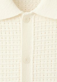 Gros plan d'une chemise en maille crème avec un motif texturé, un col pointu et deux boutons crème visibles sur la patte de boutonnage avant.