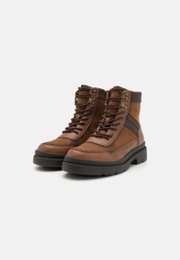 Botas de cuero marrón hasta el tobillo con superficie texturizada, ojales de metal y diseño de cordones. Presentan una suela de goma negra durable y una banda de acento lateral.
