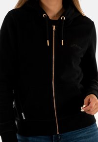 Felpa nera con zip realizzata in tessuto morbido, dotata di tasca a marsupio, zip in tono oro e cordoncini. Presenta il logo del marchio sopra la tasca.
