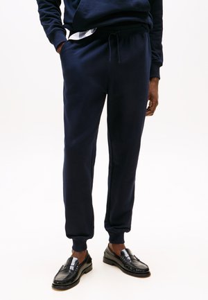 Homme portant un pantalon de jogging bleu marine avec cordon et des mocassins en cuir noir, debout devant un fond blanc.