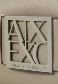 Beiger, struktureller Stoff mit einem grauen geometrischen Logo, das die Buchstaben "AX" und "EXC" zeigt, sowie dem Text "ARMANI EXCHANGE."