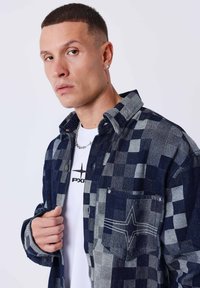 Camisa a cuadros en tonos azul marino y gris, diseño de botón, fabricada en un tejido similar al denim, con un bolsillo frontal que presenta un detalle de costura en forma de estrella.