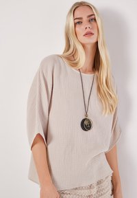 Ljust beige, strukturerad blus med lös passform och korta ärmar. Har en rund halsringning och ett lager av hängande halsband.