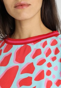 Lyhythihainen paita kevyestä sinisestä kankaasta, jossa on rohkeita punaisia ja vaaleanpunaisia abstrakteja kuvioita. Punainen ribbireuna kauluksessa tuo kontrastia ja yksityiskohtia.