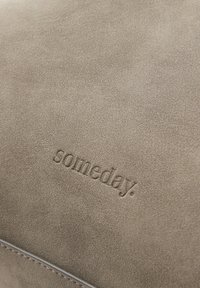 Zachte beige suède oppervlak met het woord "someday." in kleine letters geperst nabij een gestikte rand.