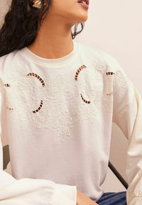 Hvid sweatshirt med blomsterbroderi og udskæringsdetaljer, lange ærmer og ribbet halsudskæring, lavet af et blødt tekstil.