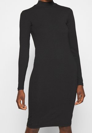 Slim-fit long-sleeve black turtleneck dress worn by a standing person with hands relaxed by their sides against a plain background.