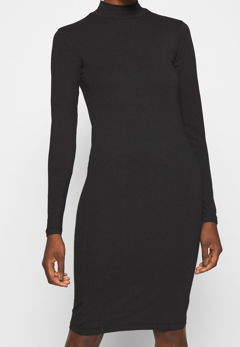 Robe noire col roulé ajustée à manches longues portée par une personne debout, les mains détendues le long du corps sur un fond uni.