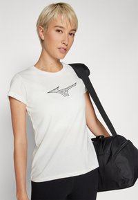 White cotton t-shirt with a short sleeve, featuring a black graphic logo on the front. A black duffel bag is slung over the shoulder.