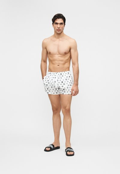 Muskuløs mann som står, iført hvite badeshorts med svarte sol- og insektmønstre, og svarte sandaler med hvit tekst.