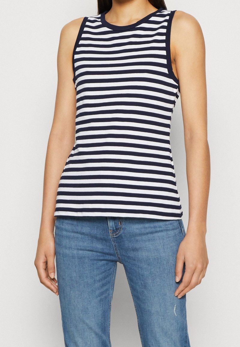 Femme portant un haut sans manches à rayures horizontales bleu marine et blanc, associé à un jean en denim bleu clair, debout avec les bras détendus.