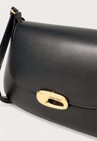 Crna kožna torba s glatkom teksturom, zlatnim ovalnim kopčanjem i remenom za rame. Ima suptilnu šavove i strukturirani dizajn.