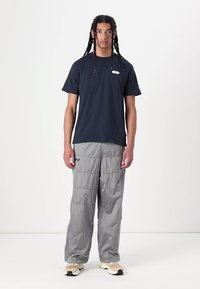 Navy short-sleeve t-shirt with a small label, paired with gray padded pants. Model stands wearing cream sneakers on a plain background.