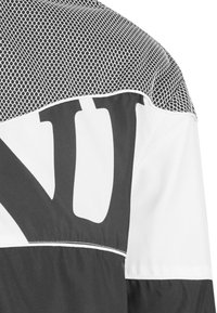 Color-blocked shirt featuring a white lower section, black upper section, and a mesh pattern at the shoulders. Large, bold black letters on front.