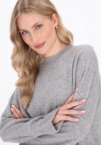 Blondynka z długimi falującymi włosami, ubrana w szary sweter, z założonymi rękami i małymi perłowymi kolczykami, delikatnie się uśmiecha na białym tle.