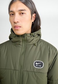 Žalias paminkštintas džemperis su užtrauktuku, reguliuojamu gobtuvu ir elastingais rankogaliais. Ant krūtinės yra juodas Nike Air logotipo užklijavimas.