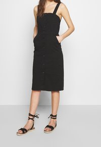 Kobieta w czarnej sukience midi na guziki bez rękawów z kieszeniami oraz czarnych płaskich sandałach sznurowanych, stojąca na tle białego tła.