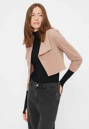 Femme aux cheveux bruns et raides portant une veste courte beige sur un top noir et un jean gris foncé, debout devant un fond blanc.
