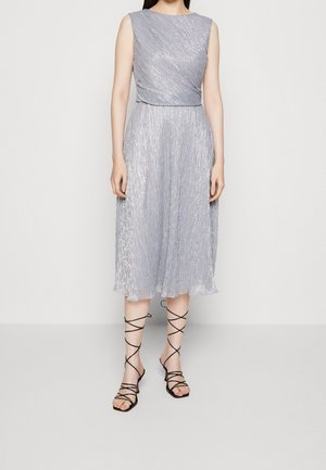 Ærmeløs sølv metallic midi kjole med folderet bodice kombineret med sorte snørede sandaler på ensfarvet baggrund.