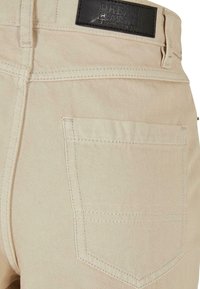 Jeans de mezclilla beige con un bolsillo trasero. Presentan una etiqueta de cuero negra con "Urban Classic" grabada. Las costuras están visibles.