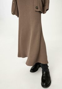 Strick, langes braunes Kleid mit Rippstruktur und seitlichen Knöpfen, kombiniert mit schwarzen Loafers und grauen strukturierten Socken, auf einem hellen Hintergrund präsentiert.