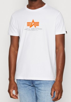 Mężczyzna ubrany w białą koszulkę z krótkim rękawem z pomarańczowym logo Alpha Industries i jasnoniebieskie dżinsy, stojący na jednolitym tle.