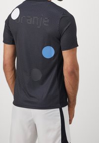 Svart idrottströja med ett prickigt mönster, stora cirkulära accentfärger i vitt, blått och grått samt textil med struktur. Designad med korta ärmar.