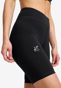 Närbild av en person som bär svarta högmidjade träningsshorts med en vit RVRC-logga på låret mot en vit bakgrund.