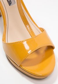 Żółte lakierowane sandały na wysokim obcasie z szerokim paskiem na palcach, otwartą konstrukcją na palce i gładkim, błyszczącym wykończeniem z detalami w postaci przeszyć.