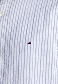 Camisa de botones a rayas en azul y blanco. Presenta líneas verticales y un pequeño logo bordado en rojo, negro y blanco en el pecho.