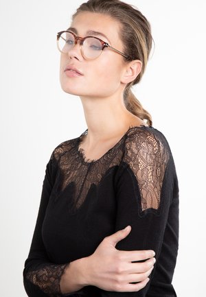 Giovane donna con capelli castano chiaro raccolti in una coda di cavallo, che indossa occhiali con montatura tartarugata e una maglia nera con spalle e maniche in pizzo, su sfondo bianco.