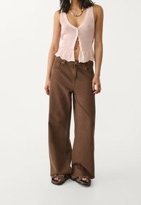 Světlounce růžová pletená vesta s rozšířeným lemem a zapínáním na knoflíky, doplněná volnými hnědými kalhotami s vysokým pasem a černými sandály.