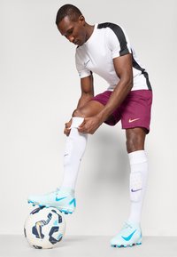 Sportovní oblečení Nike s bílým tričkem s krátkým rukávem a černými akcenty, vínovými šortkami, bílými ponožkami a světle modrými kopačkami na fotbalovém míči.