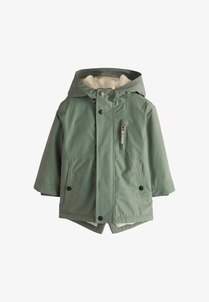 Ung dreng står indendørs iført en grøn hættetrøje, blå jeans og grønne sneakers med en beige baggrund.