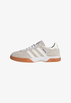 Sportliche Schuhe aus Wildleder in hellem Beige mit weißen Streifen und einer orangefarbenen Gummisohle. Sie haben eine runde Zehenform und weiße Schnürsenkel.