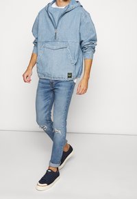 Sudadera con capucha de mezclilla azul claro con rayas texturizadas, bolsillo frontal y cierre de medio cremallera. Combinada con jeans rasgados azul claro y zapatillas oscuras.