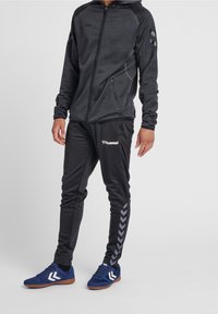 Svart hoodie med dragkedja, grå detaljer och sidfickor som matchas med svarta byxor med zigzag-mönster och logotyp. Blå träningsskor.