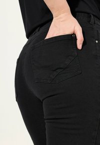 Schwarze Jeans aus Denim mit einem taillierten Schnitt und einer Gesäßtasche, die subtile Nähte aufweist. Das Material scheint langlebig und hat eine glatte Textur.