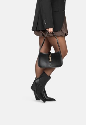 Black leather handbag with a structured shape, gold hardware, and a short strap. Paired with brown skirt and black heeled boots.