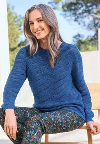 Blauer gestrickter Pullover mit strukturiertem Zickzackmuster, kombiniert mit bunten gemusterten Hosen in grünen, braunen und gelben Farbtönen.