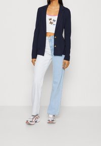 Námořnický blejzr se dvěma zlatými knoflíky, bílý crop top s motivy motýlů a světle modré kalhoty širokého střihu; model má na sobě stříbrné tenisky.