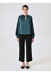 Blusa di raso in verde acqua con maniche lunghe, scollatura a chiave e dettagli arricciati. Abbinata a pantaloni neri a gamba larga e ballerine lucide a punta.