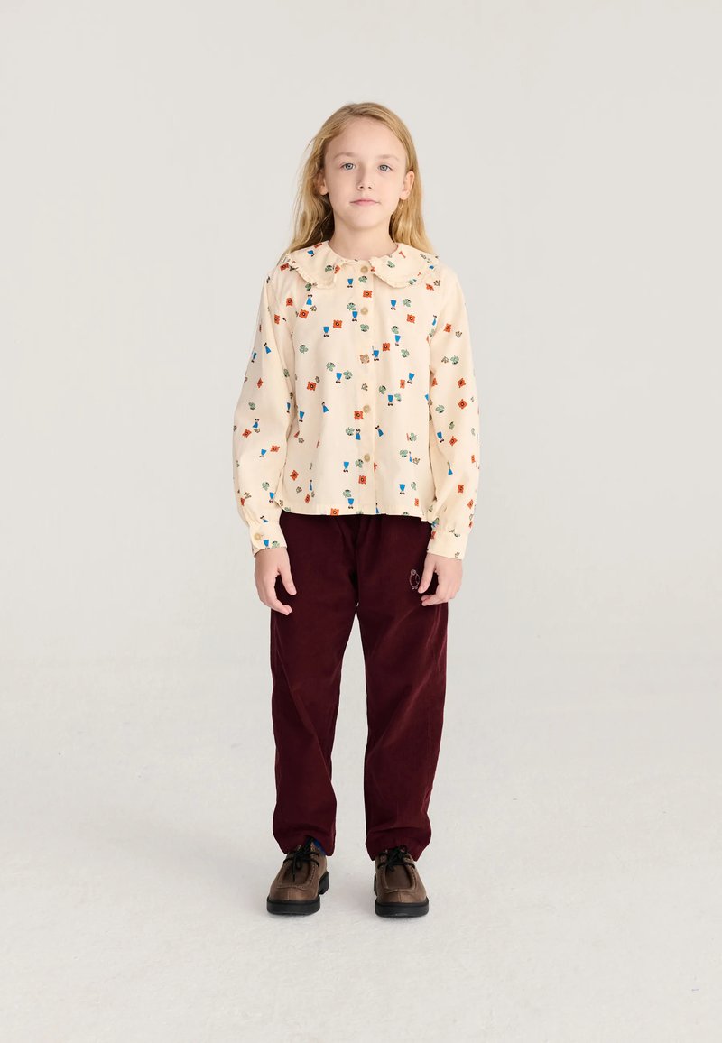 Jeune fille aux longs cheveux blonds portant un chemisier crème avec des motifs colorés, un pantalon bordeaux et des chaussures marron, debout devant un fond uni.