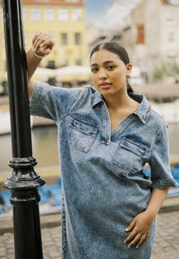 Robe chemise en denim, bleu clair avec deux poches poitrine, manches courtes et col. Le modèle pose près d'un poteau noir, à proximité de l'eau et de bâtiments.