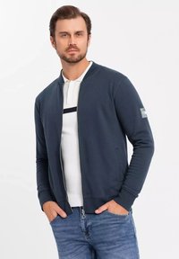Giacca bomber zip-up blu navy in morbido tessuto, con polsini a coste, tasche laterali e una patch sulla manica sinistra. Indossata su un polo bianca.